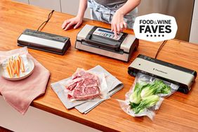 Person pressing control panel on one of three vacuum sealers on wooden countertop with different vacuum-sealed bags of food