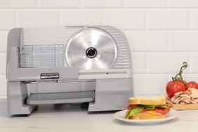 Chef'sChoice 615A Electric Meat Slicer For Home Use on a white surface next to a sandwich, cold cuts, and tomatoes 