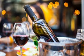 A glass of red wine with a bottle of red wine in an ice bucket.